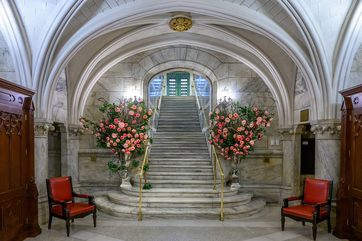 The sacristy staircase: a broad marble stair rising under a vaulted arch to the bronze doors at cathedral level, flanked by tall arrangements of roses.