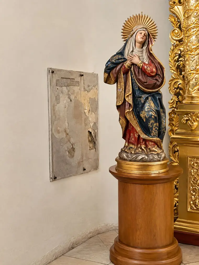 A polychrome Virgin with a golden sunburst halo and hands crossed at her breast, on a wooden pedestal beside the gilded retablo.