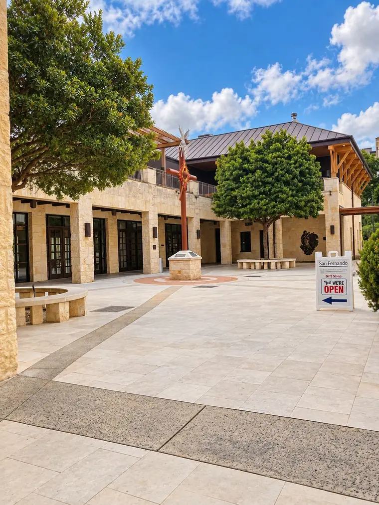 Plaza de la Cruz: a paved courtyard with a tall red cross on a stone plinth at its center, the Cathedral bookstore arcade along the left, and a sandwich board reading San Fernando Gift Shop, Yes We're Open.